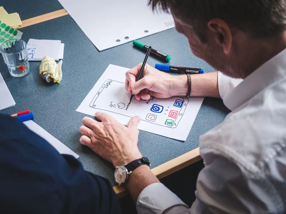 A designer sketching a mobile app screen layout on paper at a desk, illustrative of planning an icon set before it ships